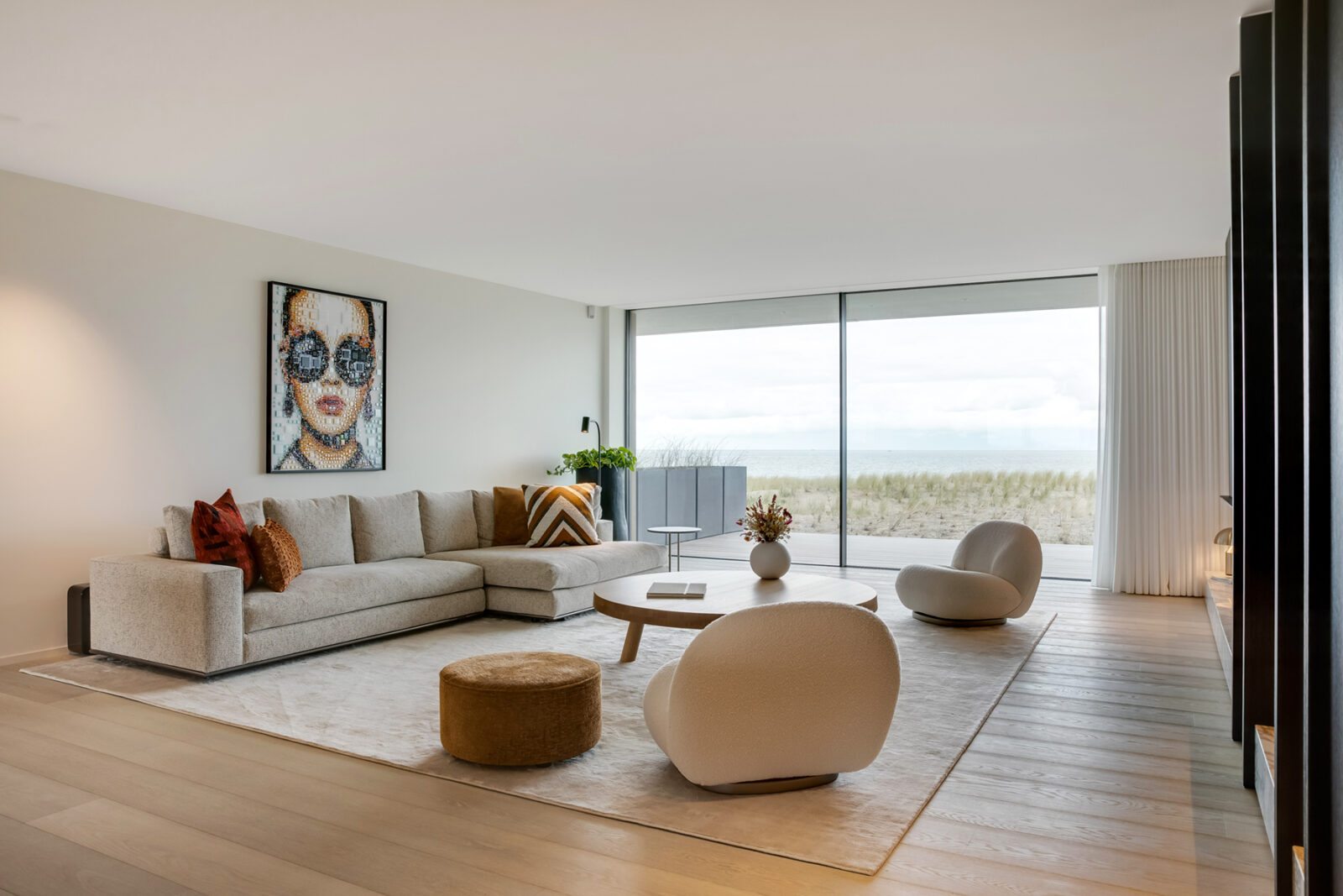 Lichte woonkamer met ronde fauteuils, een hoekbank en een lage houten salontafel, gericht op een glazen wand met uitzicht op de zee.