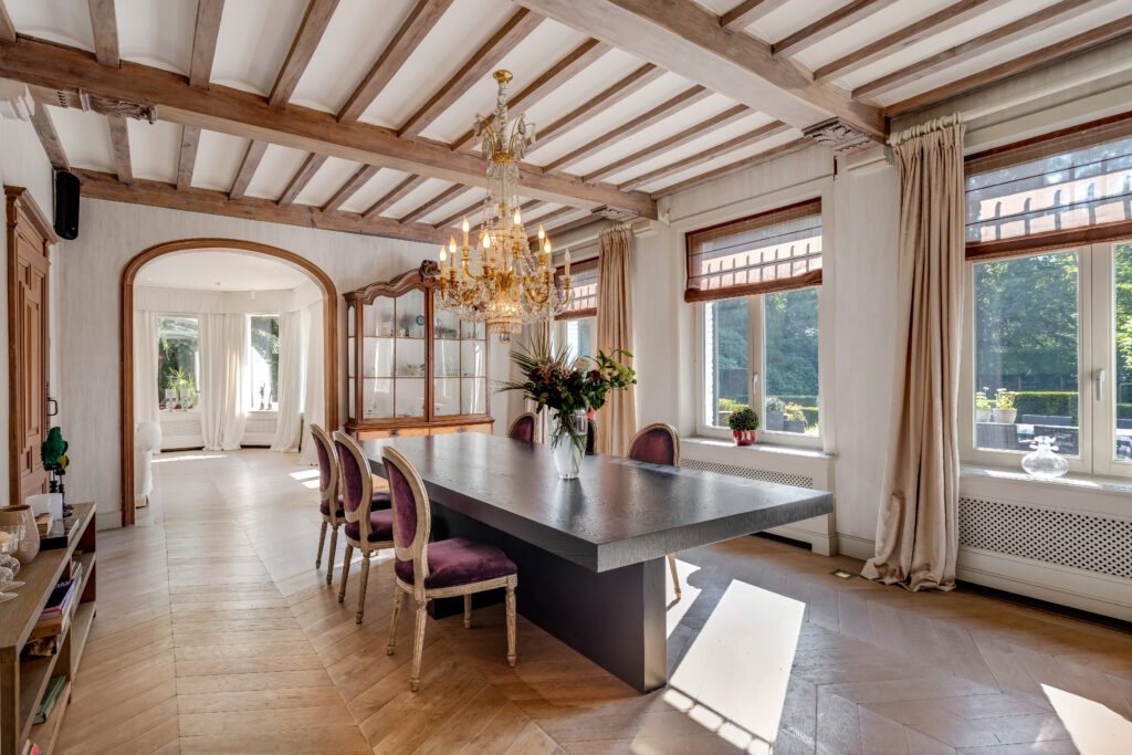 Ruime eetkamer met houten plafondbalken, kroonluchter en klassieke eettafel met fluwelen stoelen.