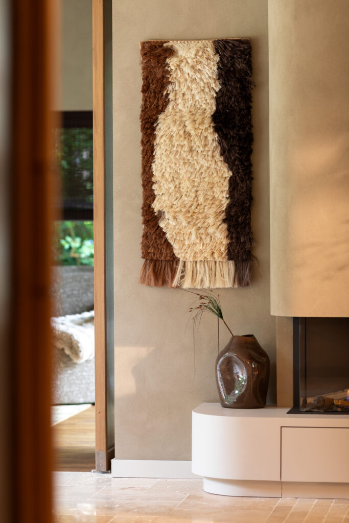 Detail van een interieur met een handgemaakt wandtapijt in bruine en creamtinten, een keramische vaas met takje en een afgeronde haard met mortexafwerking.