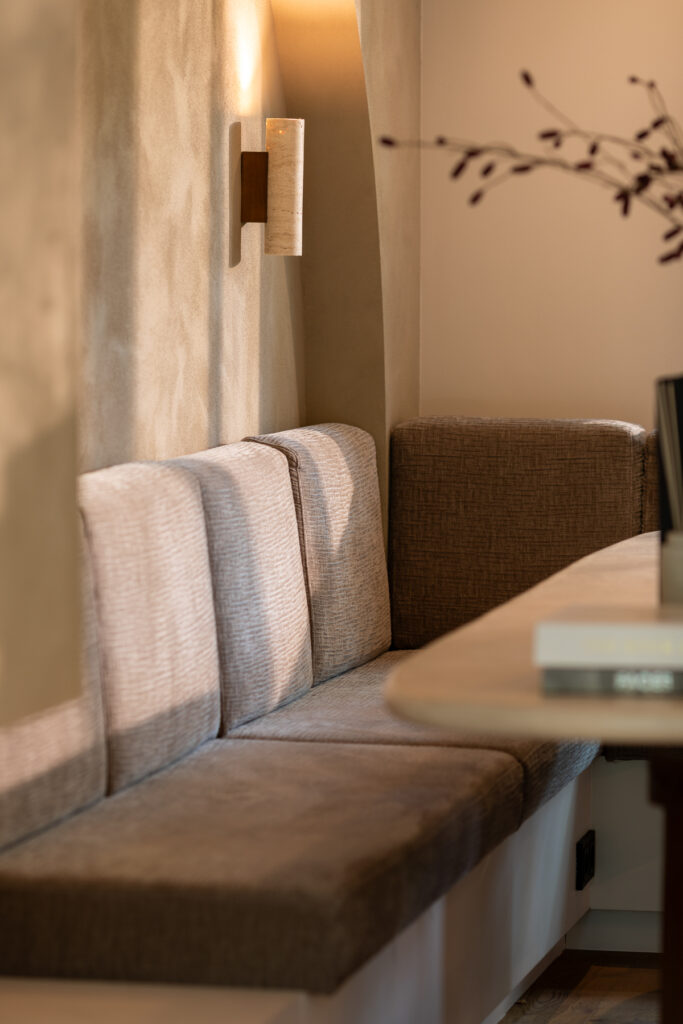 Detailfoto van een ingebouwde zithoek met taupekleurige stoffering, warme natuurlijke lichtval op een mortexwand en een minimalistische wandlamp.