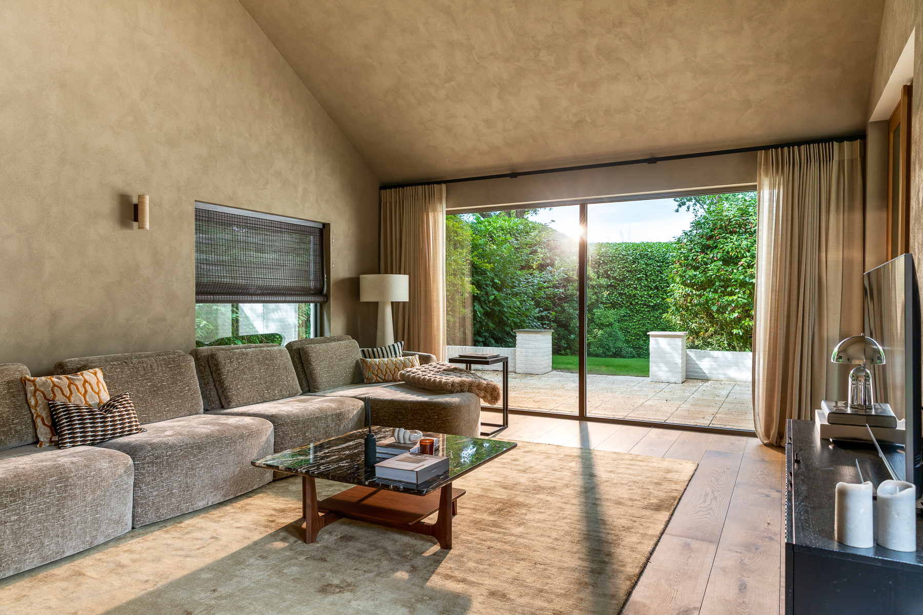 Warme woonkamer in neutrale tinten met een grote hoekbank, houten salontafel en hoge schuifpui naar de groene tuin. Zachte beige wanden en natuurlijke materialen zorgen voor een rustige sfeer. De foto benadrukt de verbinding tussen de leefruimte en het terras buiten.