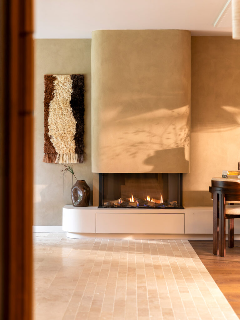 Moderne open haard met een afgeronde stucwerk schouw en lage ingebouwde haardpartij tegen een warme beige wand. De lichte tegelvloer en het vuur zorgen voor extra warmte in het interieur. De compositie legt de nadruk op de haard als sculpturaal middelpunt van de ruimte.