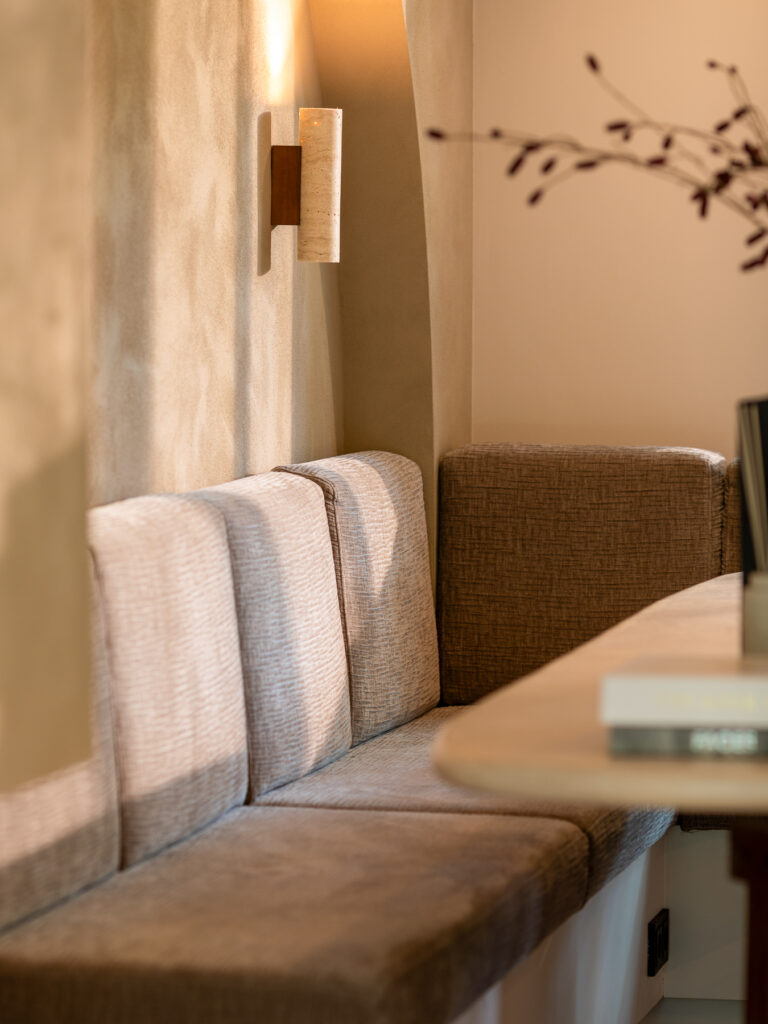 Close up van een gestoffeerde vaste bank langs de wand met een minimalistische wandlamp erboven. De warme wandafwerking en zachte stoffering zorgen voor een rustige uitstraling. De afbeelding legt de focus op materiaal, textuur en sfeerverlichting in de ruimte.