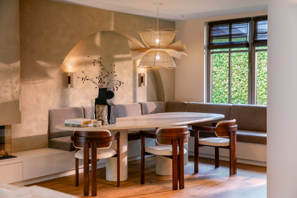 Maatwerk vaste bank rond een ovale eettafel gecombineerd met massief houten stoelen. Een subtiele boog in de wand en een geweven hanglamp geven de ruimte karakter. De afbeelding laat zien hoe comfort en natuurlijke materialen samenkomen in deze eethoek.