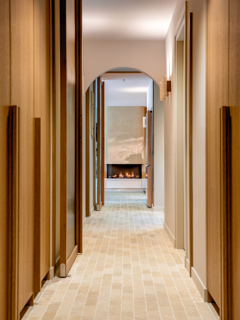 Smalle gang met een ronde boogdoorgang en lichte natuurstenen vloer. Zacht wandlicht en houten deuren zorgen voor een warme en rustige uitstraling. De foto legt de nadruk op symmetrie en diepte in de architectuur.