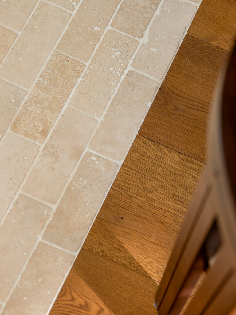 Detailfoto van de overgang tussen een lichte natuurstenen tegelvloer en een warme houten vloer. De verschillende materialen sluiten strak op elkaar aan. De afbeelding toont de zorgvuldige afwerking en het contrast in texturen binnen het interieur.