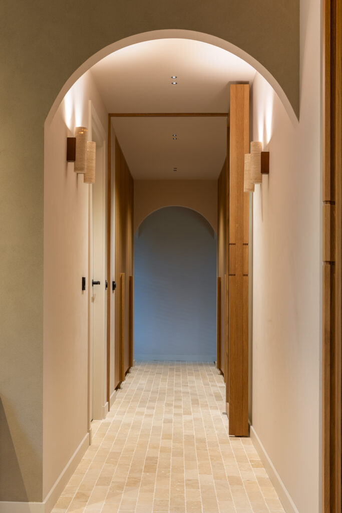 Lange gang met herhalende ronde doorgangen en een lichte natuurstenen tegelvloer. Warme wandverlichting en houten kozijnen zorgen voor diepte en ritme in de ruimte. De foto benadrukt de architectonische lijnen en het materiaalgebruik.