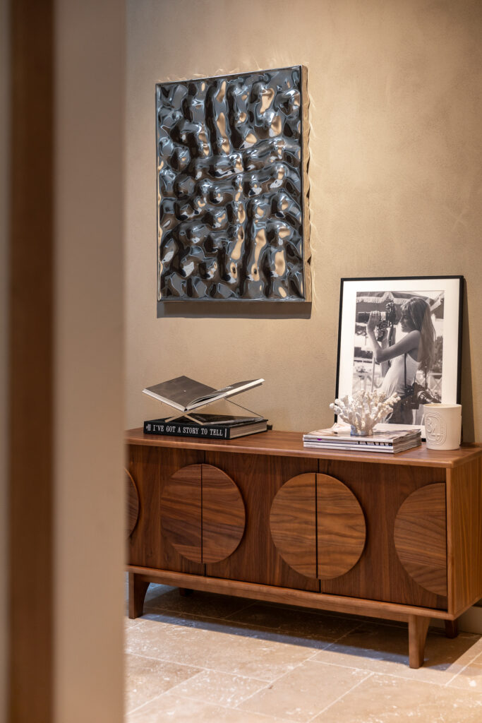 Houten vintage dressoir met ronde vormen, gestyled met boeken, een kunstobject en een zwart wit foto aan de muur. Boven het meubel hangt een reliëf kunstwerk in donkere tinten. De afbeelding toont hoe styling en karakter samenkomen in het interieur.