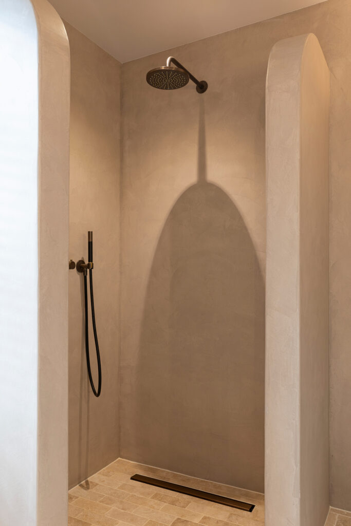 Minimalistische inloopdouche met een ronde nis en wanden in warme microcement afwerking. Een donkere regendouche contrasteert met de lichte natuurstenen vloer. De afbeelding benadrukt de rustige en spa achtige uitstraling van de badkamer.