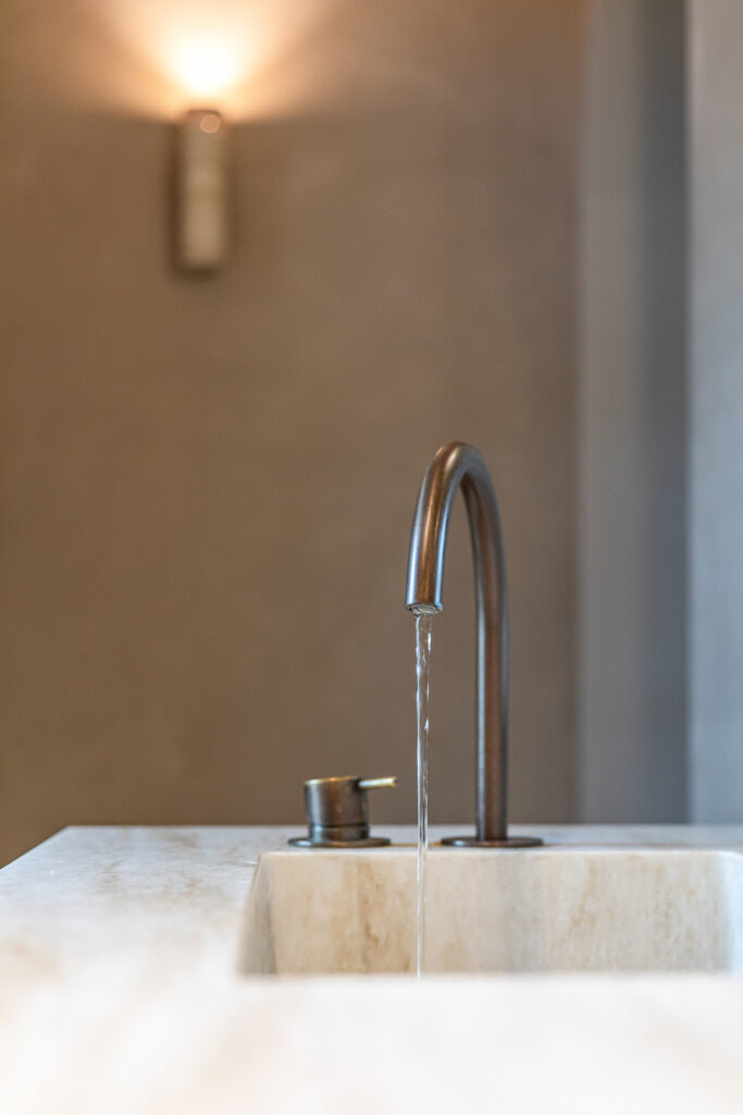 Rechthoekige natuurstenen wastafel met een slanke wandgemonteerde kraan tegen een warme beige wand. Subtiele wandverlichting zorgt voor een zachte gloed boven de wasbak. De foto toont een verfijnd detail binnen het badkamerontwerp.