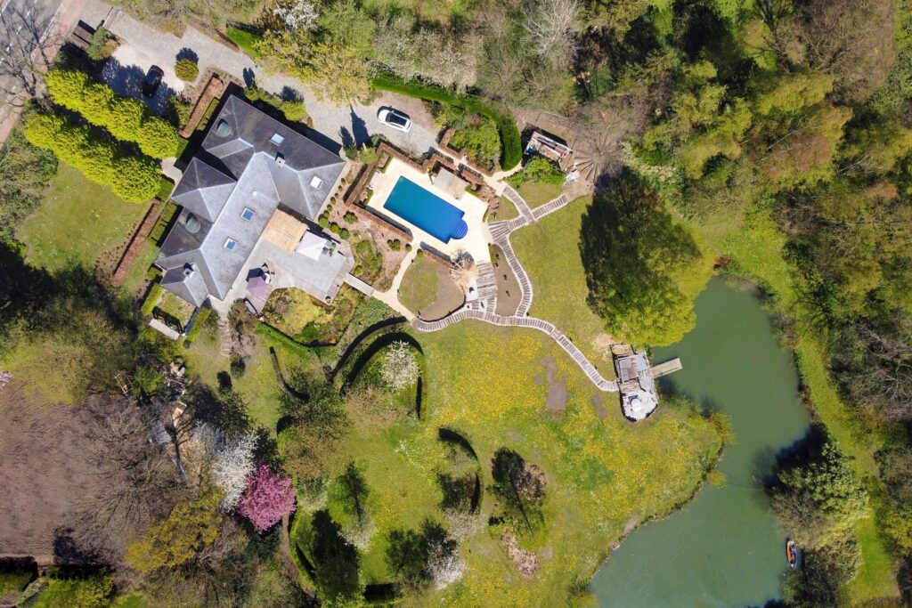 Dronefoto van een bovenaanzicht van een woning gelegen is een bosrijke omgeving, voorzien van een zwembad, een grote vijver en een enorme oprijlaan. De foto geeft context over het perceel dat bij de woning hoort wat nooit gemaakt zou kunnen worden met foto's vanaf de grond. 